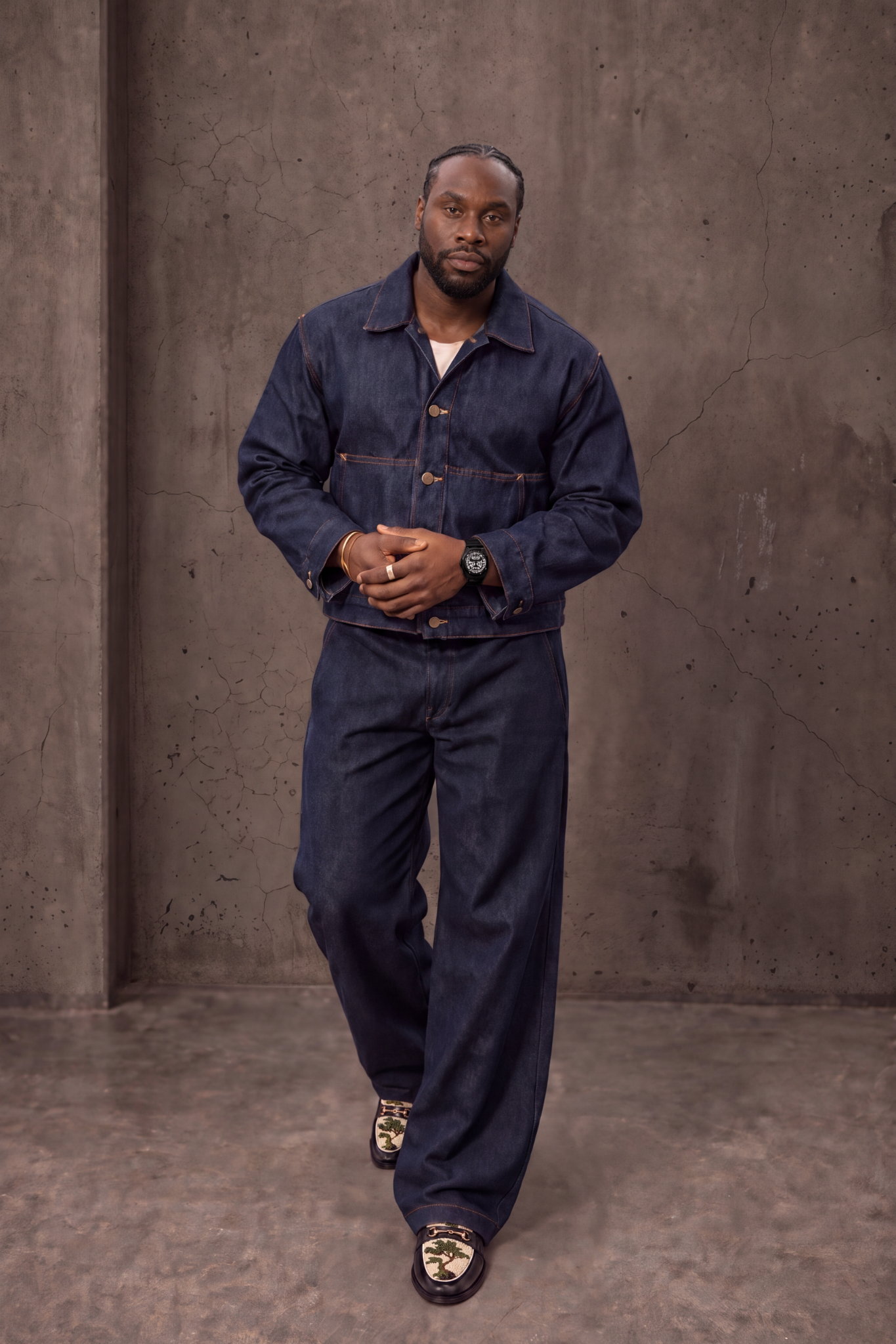 Charles Assa en pied, mains jointes, en tenue bleu indigo et mocassins à motif.